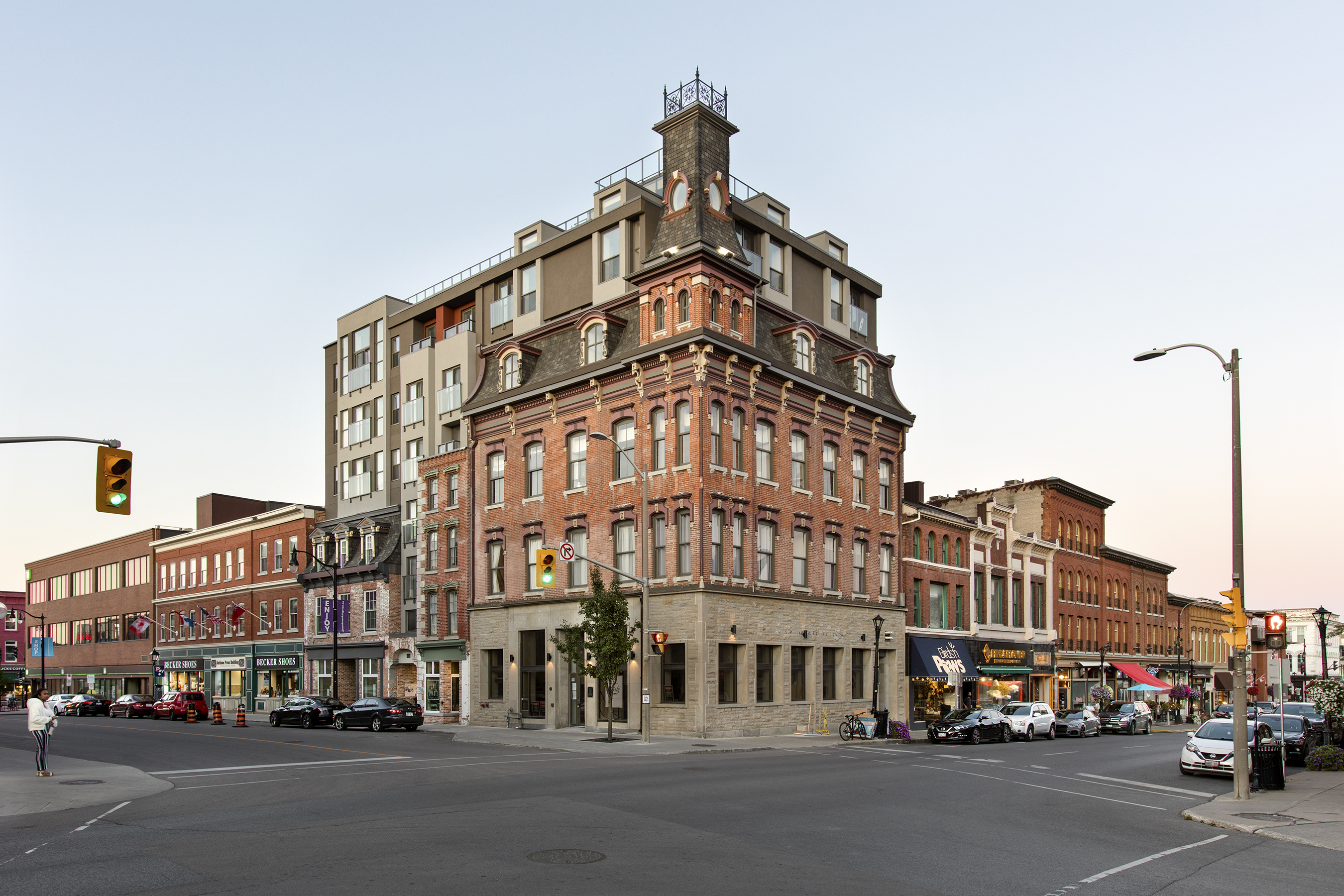    	 Street view of a historic brick building on a corner, featuring ornate architectural details and a mansard roof with dormer windows. A modern multi-story extension rises above the original structure. The surrounding streetscape includes a row of smaller brick buildings housing shops and restaurants.